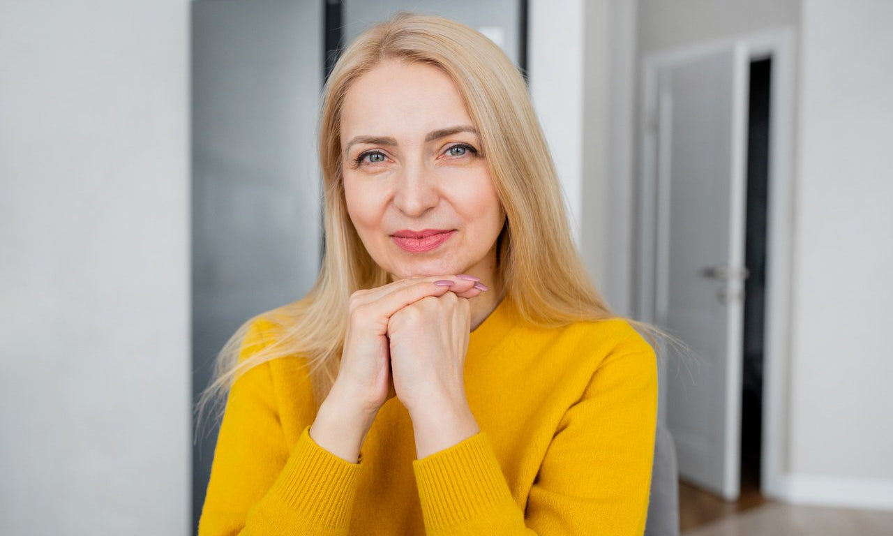Smiling middle-aged woman in a yellow sweater resting her chin on her hands, showing soft, natural lip color indoors