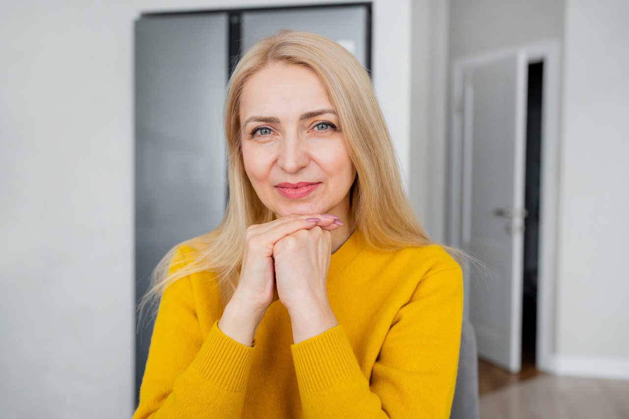 Smiling middle-aged woman in a yellow sweater resting her chin on her hands, showing soft, natural lip color indoors