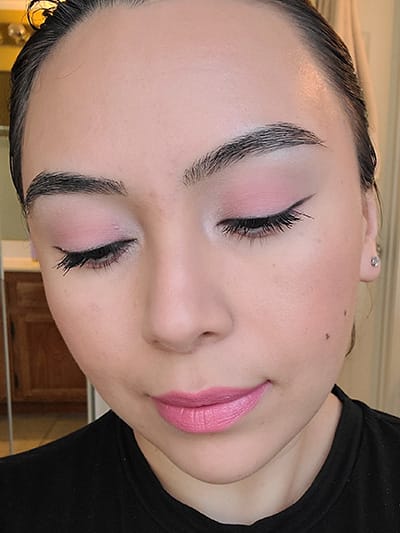 Image of female with light to medium skin tone, Black Hair, Dark eyebrows and dark brown eyes. Pink shades for eyeshadows, Pink shades of blush on her cheeks and Mauve Me Lipstick. She is looking straight on but eyes downward to show off the beautiful pinks in her monochromatic makeup look