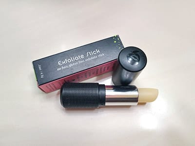 Image of Exfoliate Stick in it's Red Apple Lipstick package and Rallye Balm with the bullet twisted up and the cap laying next to it on a table top