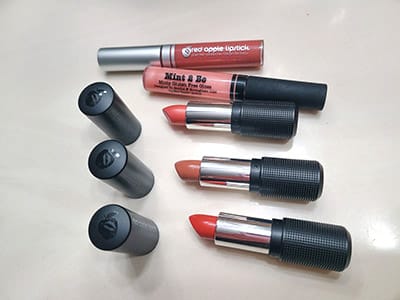 Image of an assortment of 3 Red Apple Lipstick lipstick tubes with the bullet with the caps lined up at the tops of the tubes along with 2 Lip Glosses by Red Apple lipstick.