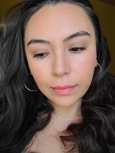 Image of lady with medium skin tone, long dark hair and dark eyes. She is wearing a  light makeup application of Prime Time primer on the lids, Heirloom, Blondeshell, and Buttercream eyeshadows and The Lash Project mascara on the eyes; Sundrop Bronzer and Tango Blush; wearing Coral Crush lipstick as a lip stain. All products featured are from Red Apple Lipstick INC