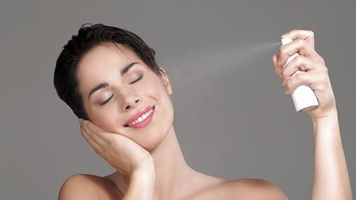 Image of stock model showing herself spraying a facial mist onto her face with her head tilted to the right and her cheek in the opposite palm of her hand